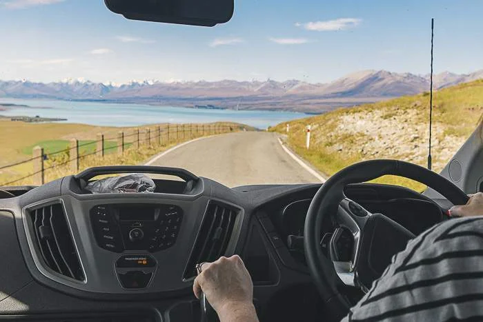 Driver driving a cheap campervan hire New Zealand with view of a lake and mountains through the windscreen